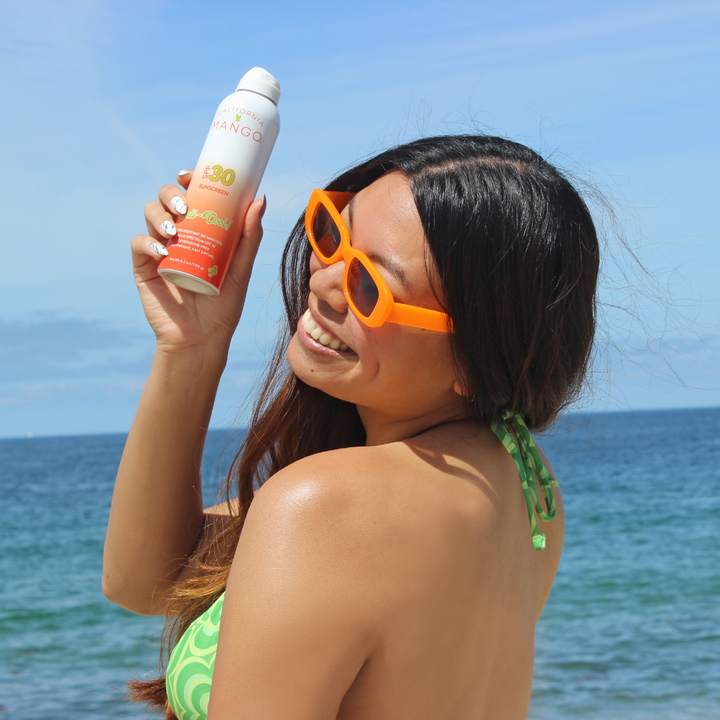 California Mango SPF 30 sunscreen spray bottle held up at a sunny beach.