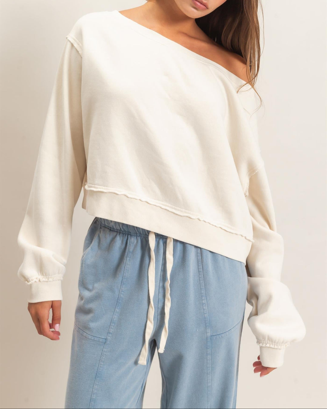 HYFVE Raw Edge Off the Shoulder Sweatshirt in Alpaca cream color, paired with blue joggers