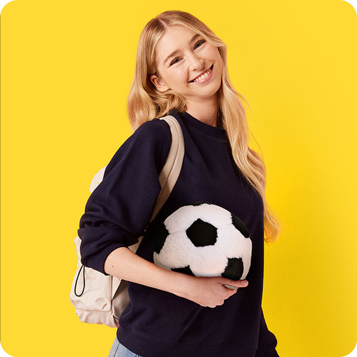 Woman holding Warmies soccer ball plush toy against yellow background